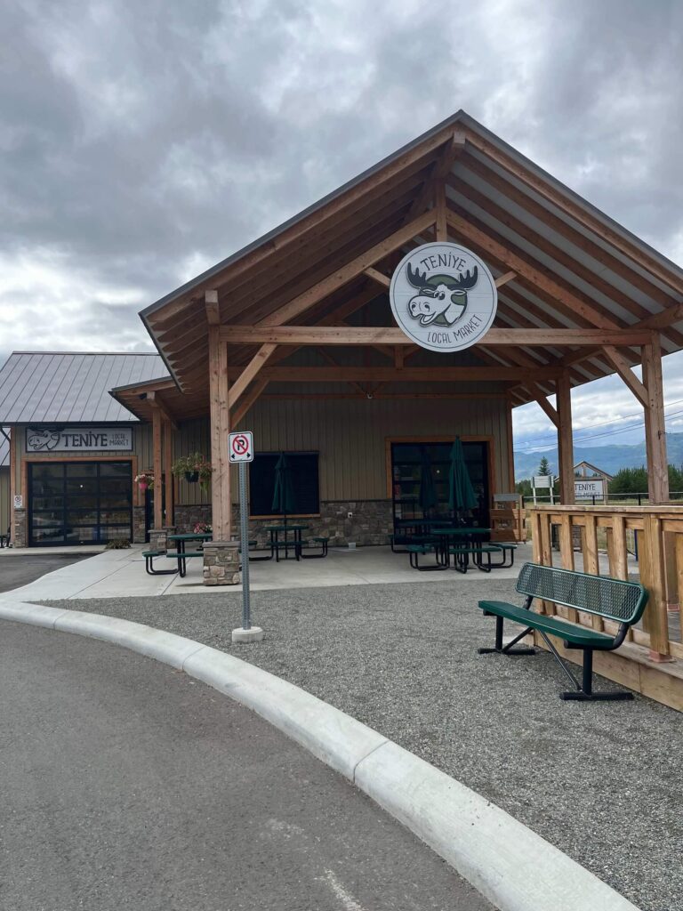 Wood-framed building of Teniye Local Market with picnic tables and mountain sky backdrop in Barriere, BC.