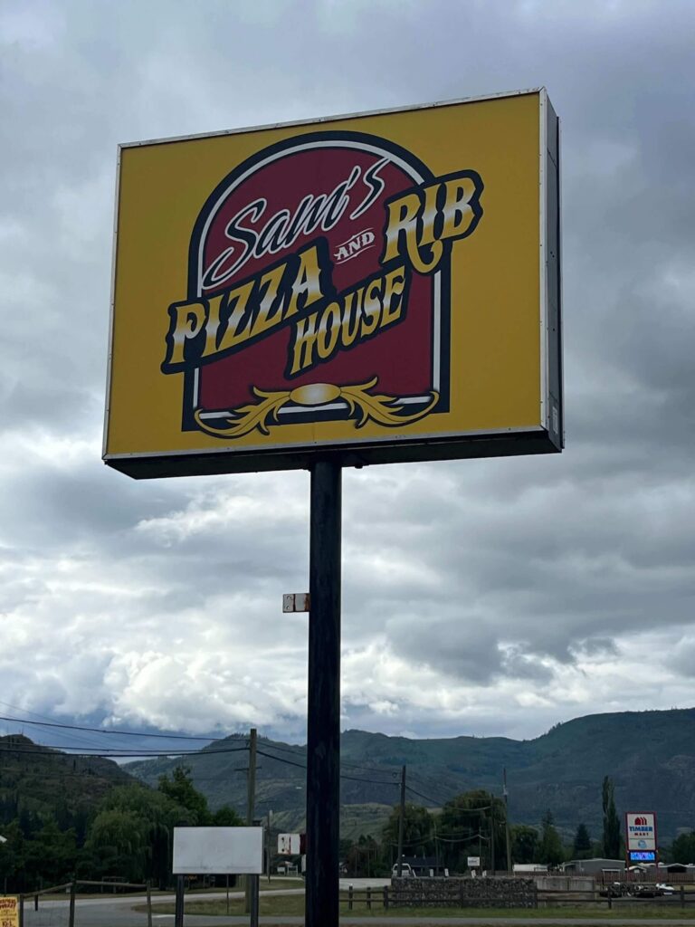 Tall roadside sign for Sam’s Pizza and Rib House with mountains and cloudy sky in the background, Barriere, BC.